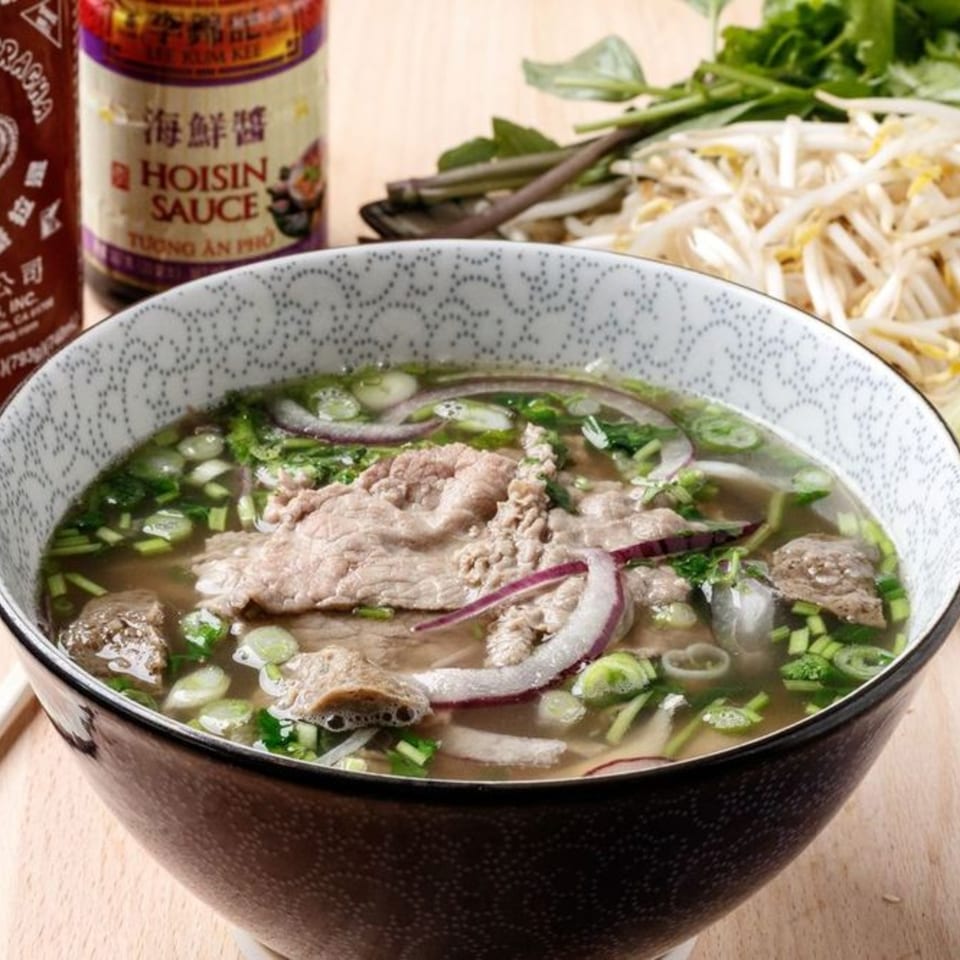 Vietnamese pho with beef, onions, and scallions in a patterned bowl, with hoisin sauce and bean sprouts.